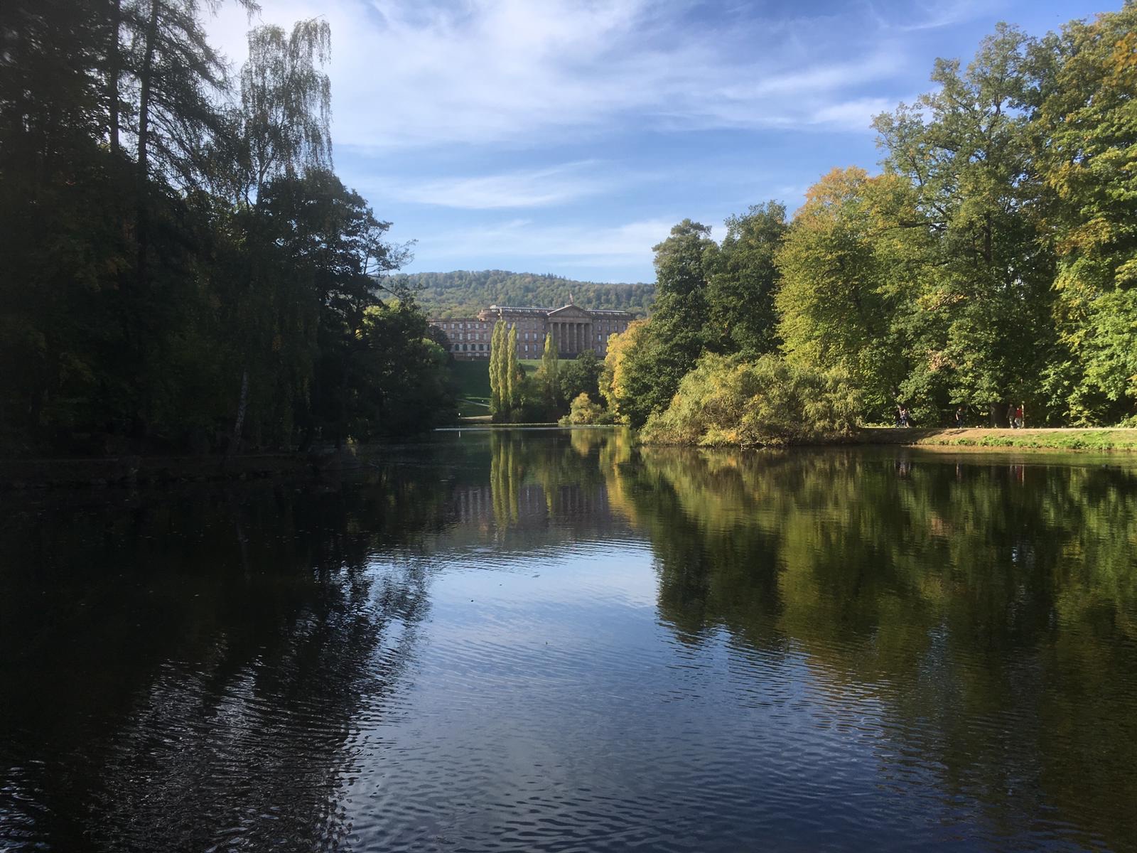 Von der Sonne beschienene Bäume und ein Schloss im Hintergrund spiegeln sich im See.